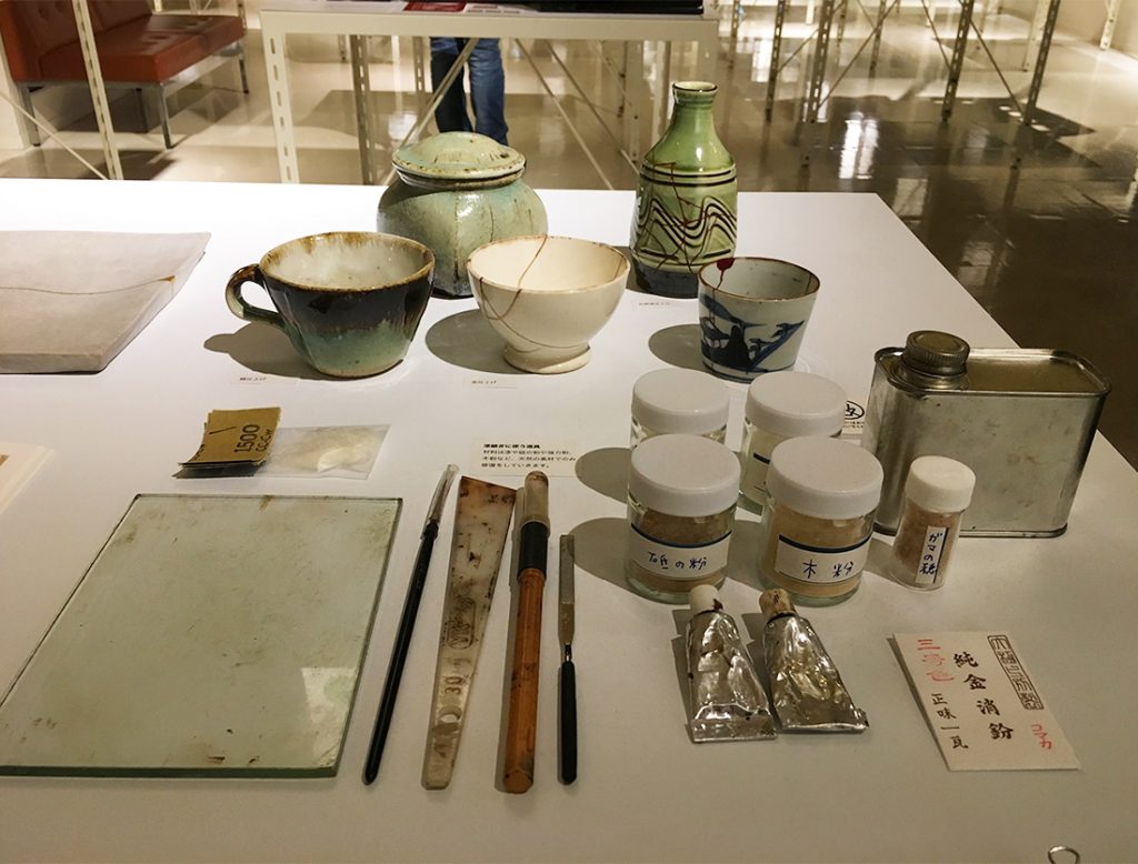 漆継ぎに使う漆や砥の粉や小麦粉などが並ぶ岩手県の展示スペース。桑原さんは「天然の材料だけで継いでるんですね」と興味津々。 sIMG_1925.atari_