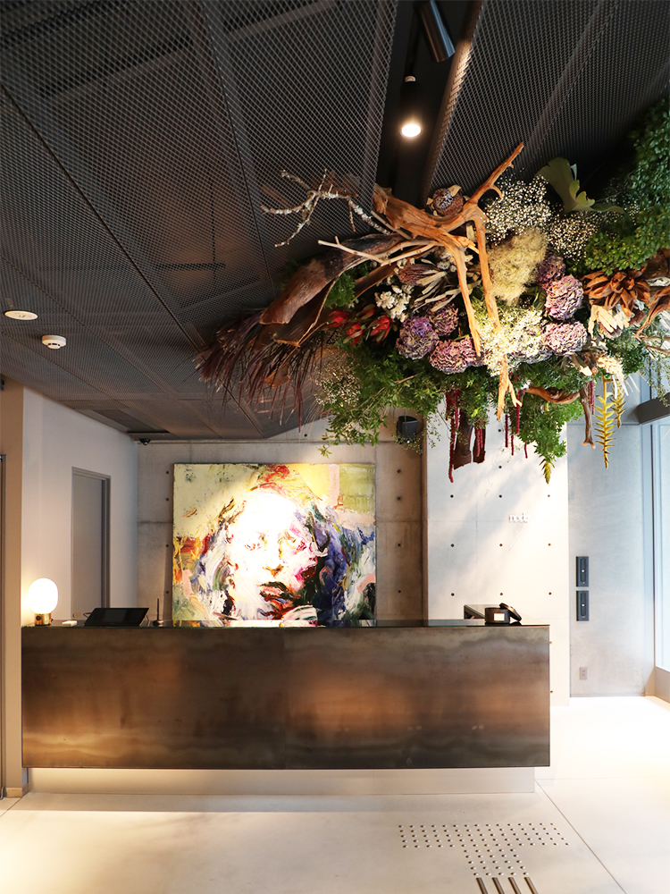 グレートーンに統一されたコンクリートの空間に、作品が映えるエントランス。中目黒の花屋〈farver〉が手がけた植物のインスタレーションも天井を有機的に彩る。