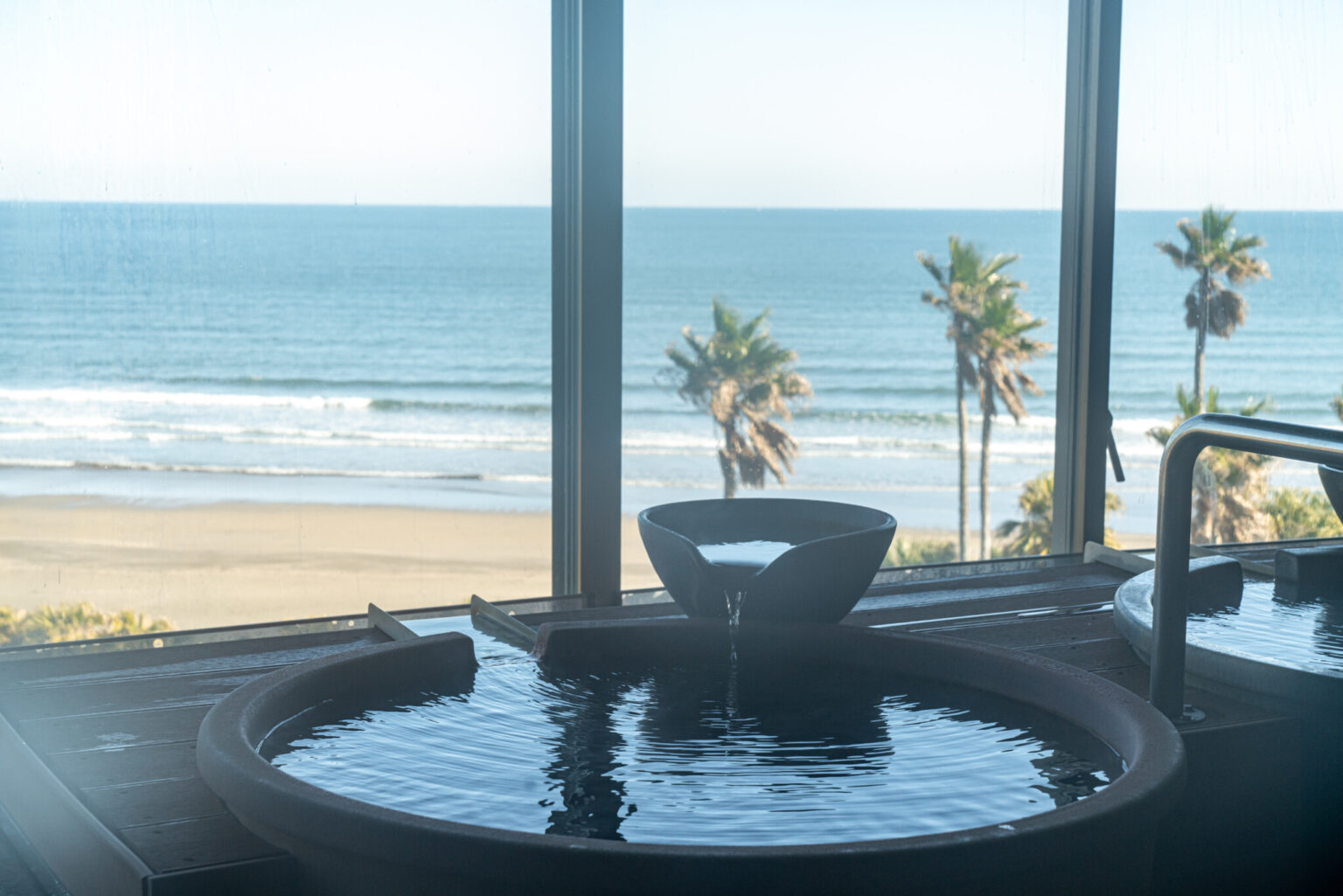 女風呂からの景色。美しい砂浜と青い海、パームツリーを眺めながらの入浴は、なかなかできない体験だ。温泉の日帰り利用は大人1,100円（税込）。『トロピカルな景色と温泉の不思議なコラボ！』