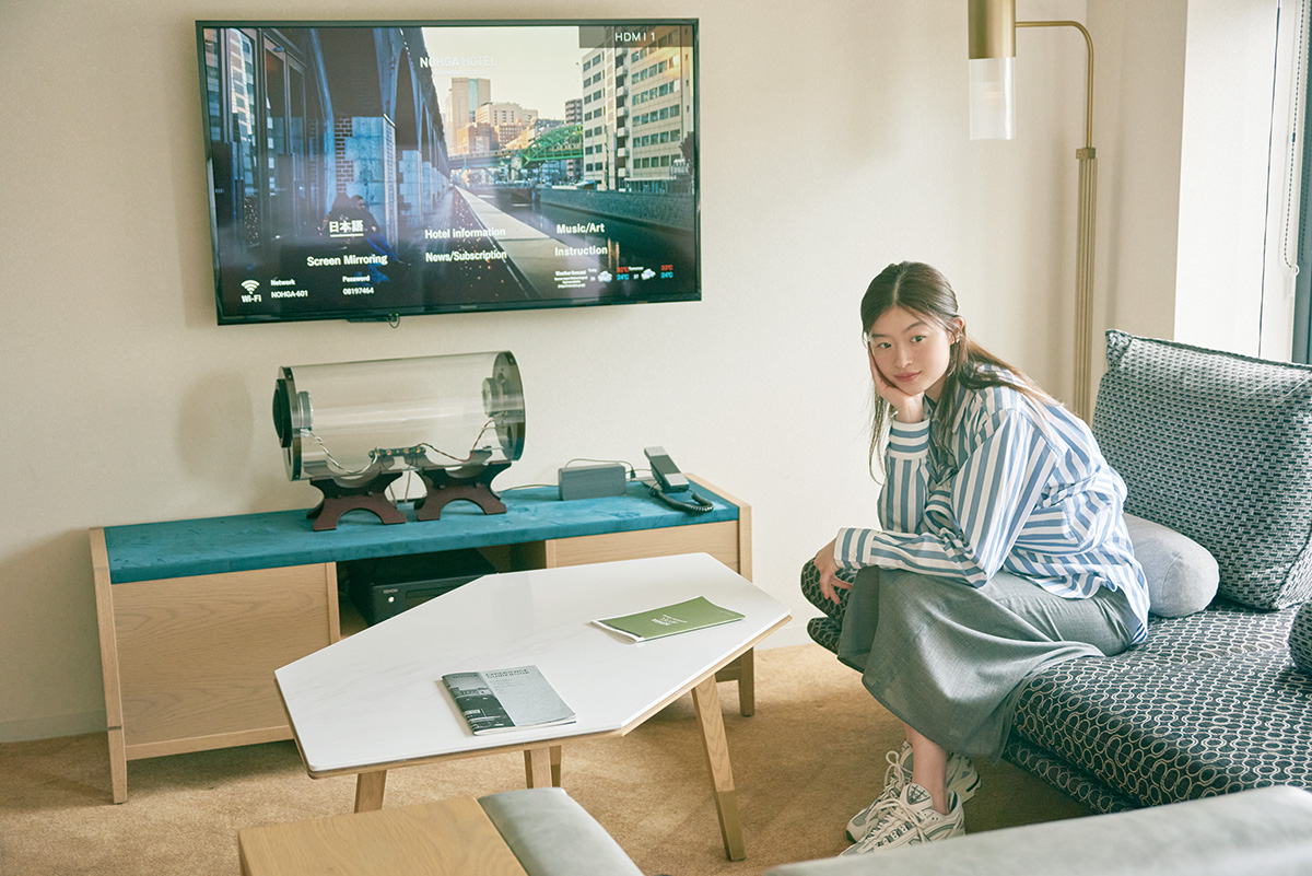 透明な筒形の〈M’s System〉のスピーカーで音楽が楽しめる部屋。ストライプ2in1シャツ64,900円（サイ｜マスターピースショールーム 03-6407-0117）／スカート49,500円（エイトン｜エイトン青山）／スニーカー19,800円（ニューバランス｜ニューバランスジャパンお客様相談室 0120-85-7120）