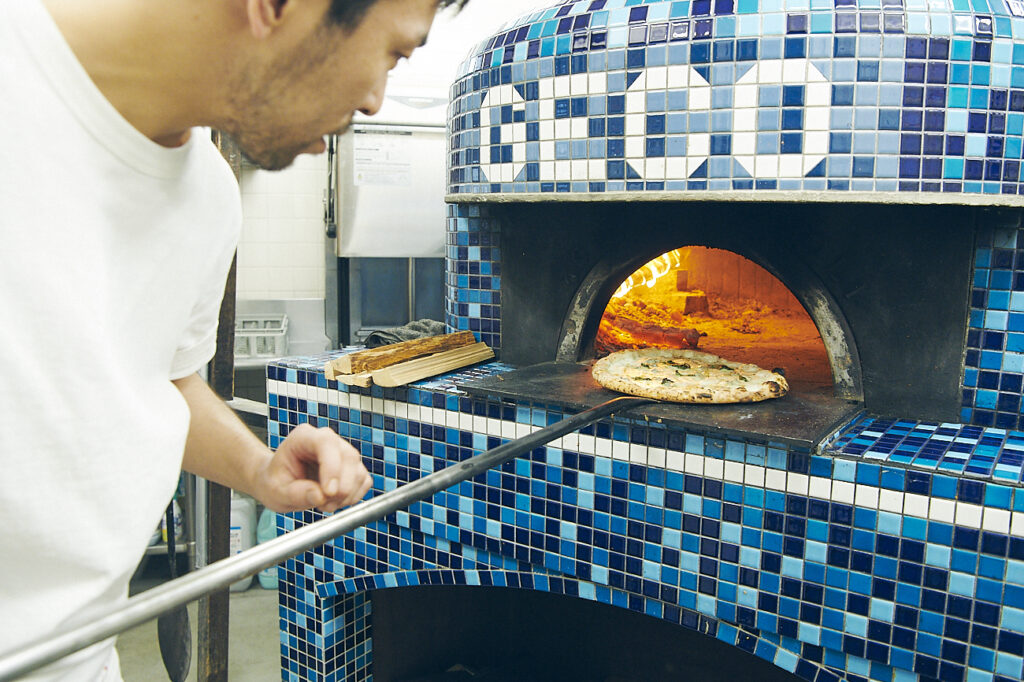 店名の入ったオリジナルの薪窯でピッツァを焼く栗原さん。1〜2分で火が入ってしまうピッツァを同時に3枚も焼ける技をもっている。